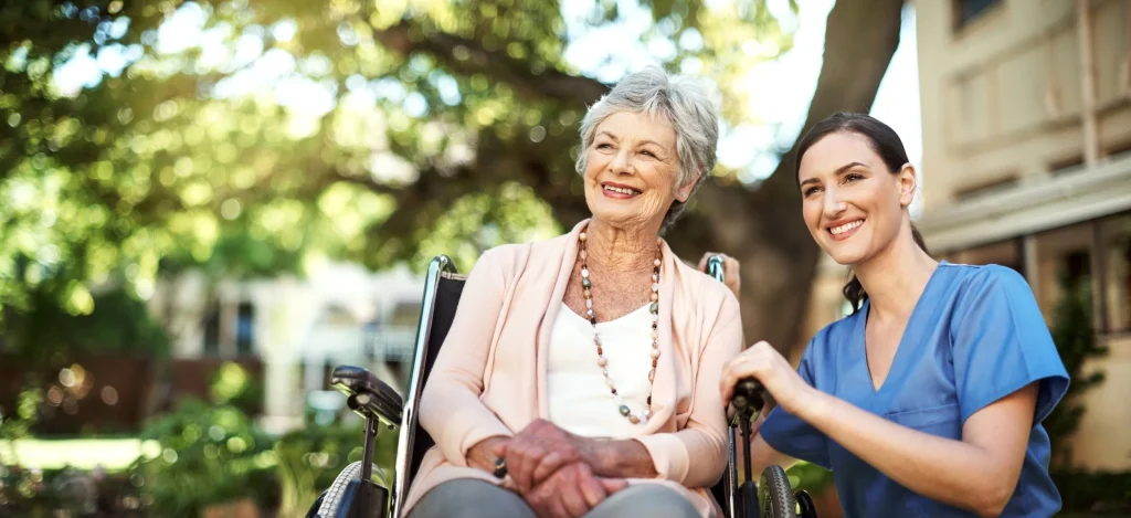 An elderly woman in a wheelchair smiling and holding hands with a young nurse in blue scrubs outdoors in a park.