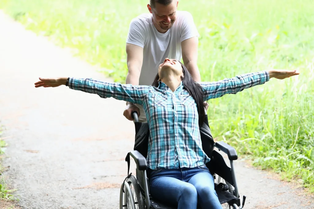 A woman in a wheelchair with arms outstretched, being pushed on a nature trail by a smiling man.