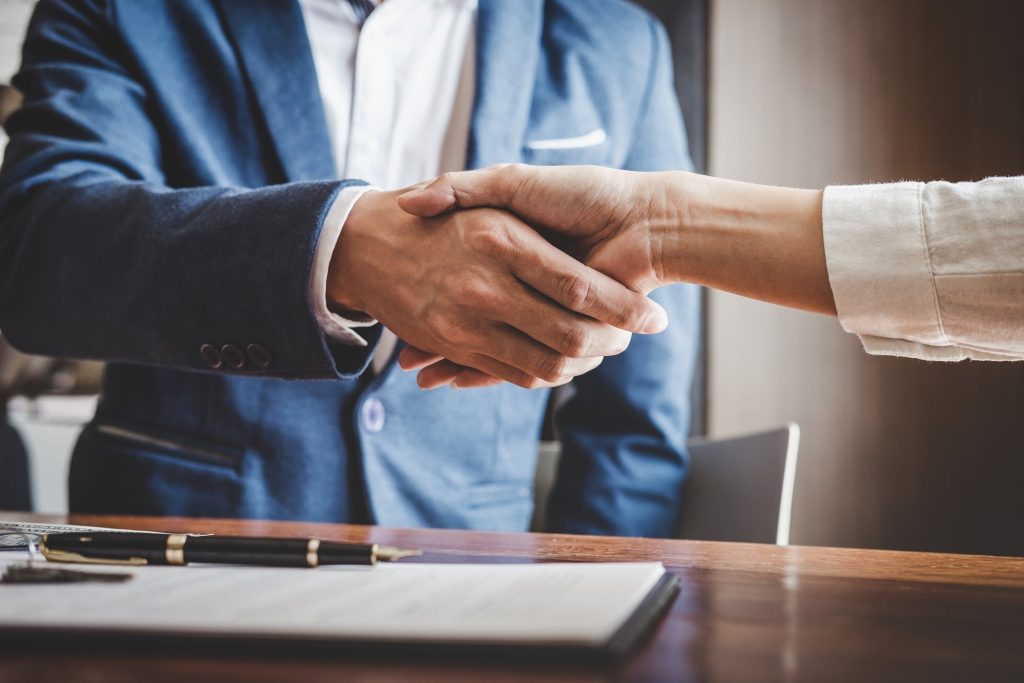 Two people shaking hands in a professional setting, with a desk and pen in the foreground.